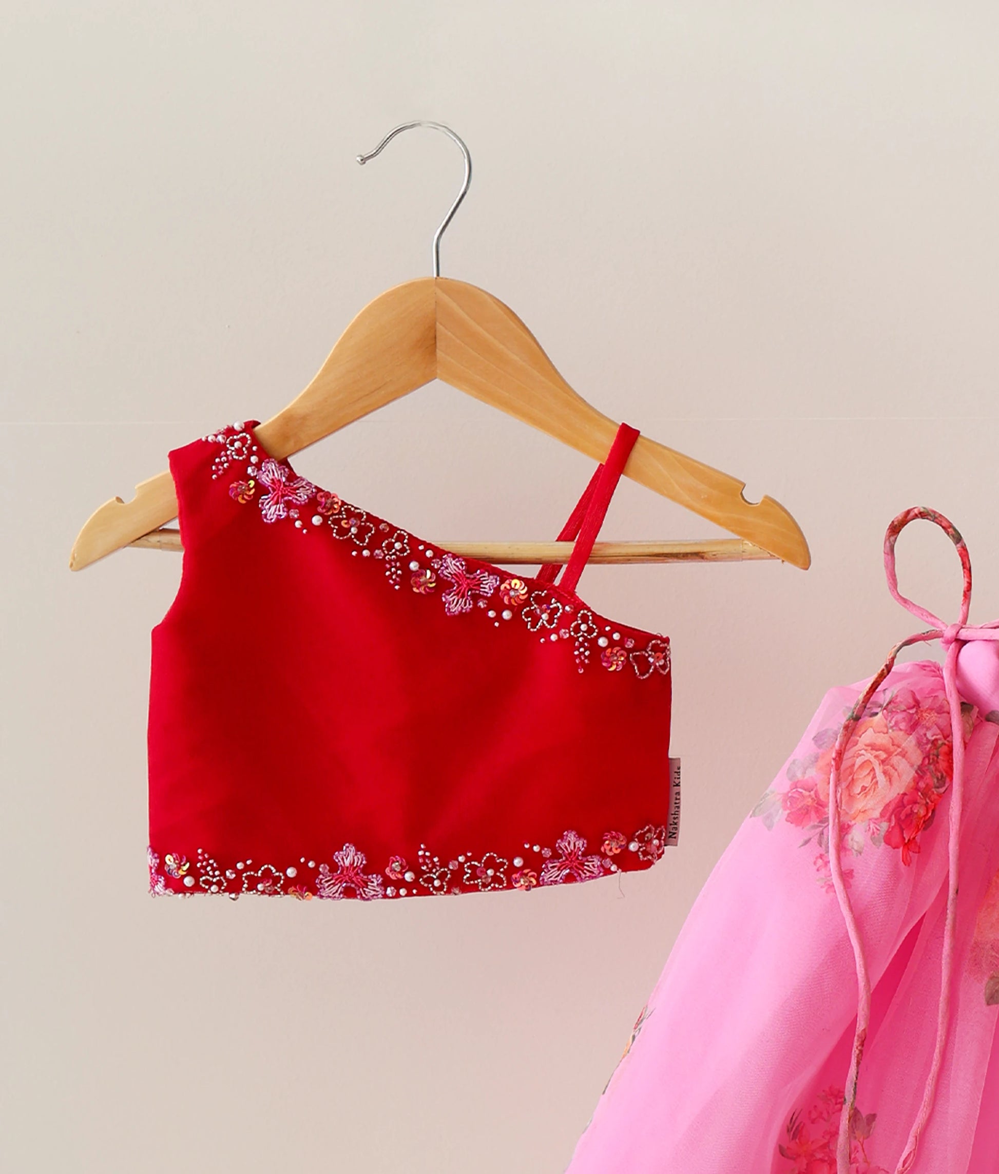 Close view of a red and pink kids' lehenga set. One-shoulder top with handwork. Pink skirt with floral prints