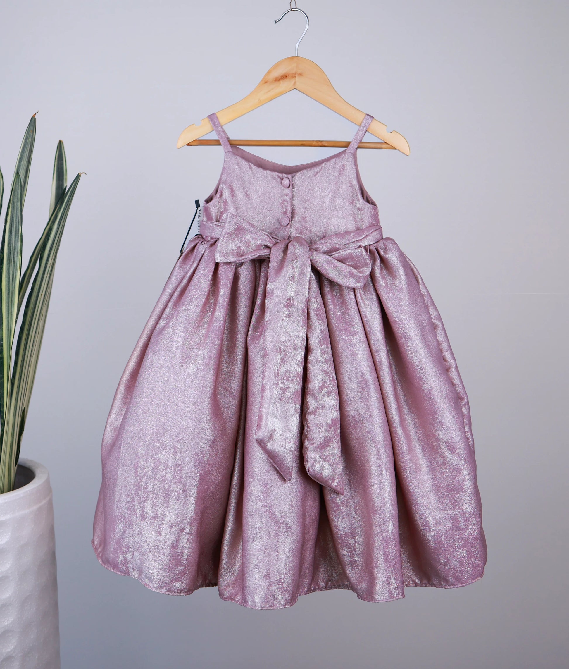 Back view of an onion pink dress with a bow on a wooden hanger against a plain background.