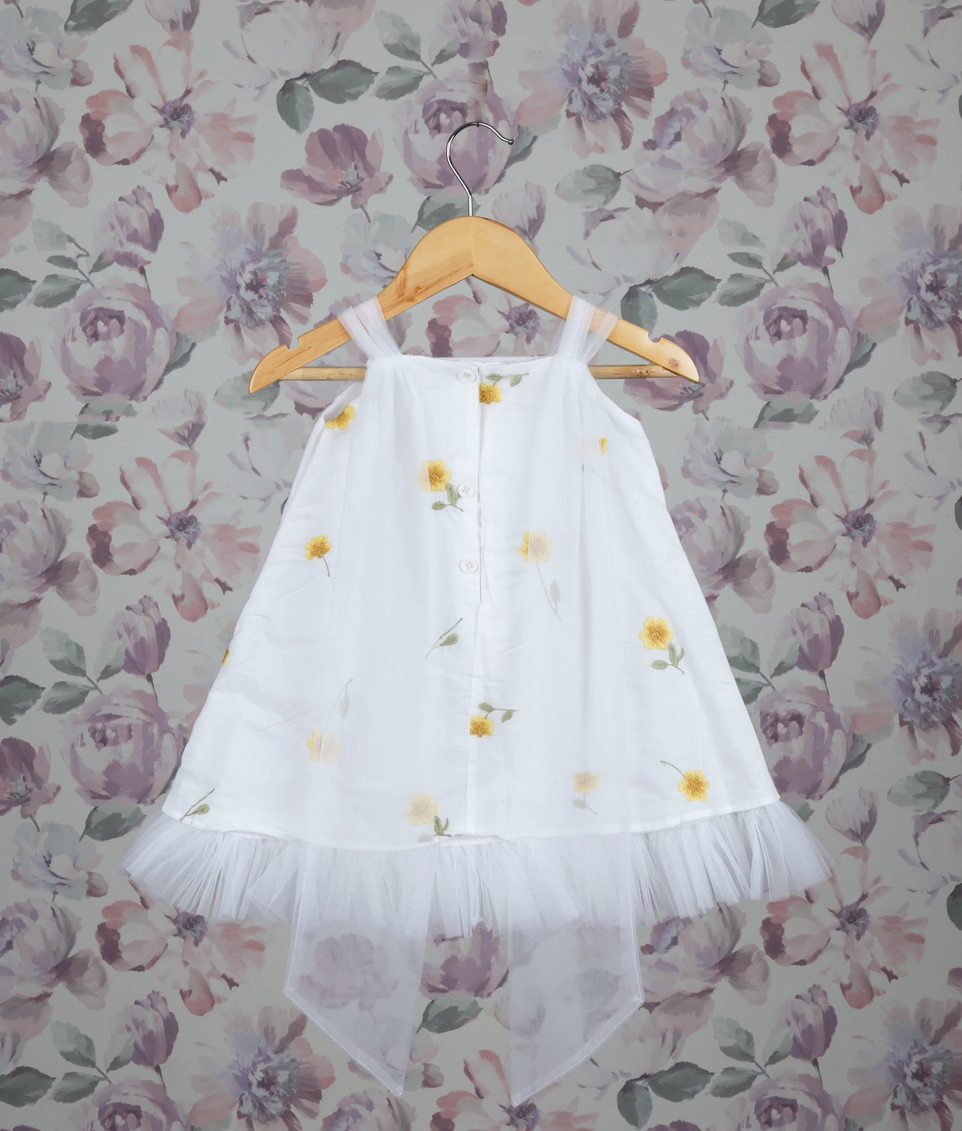 Back view of a white dress with yellow floral patterns against a floral wallpaper background.