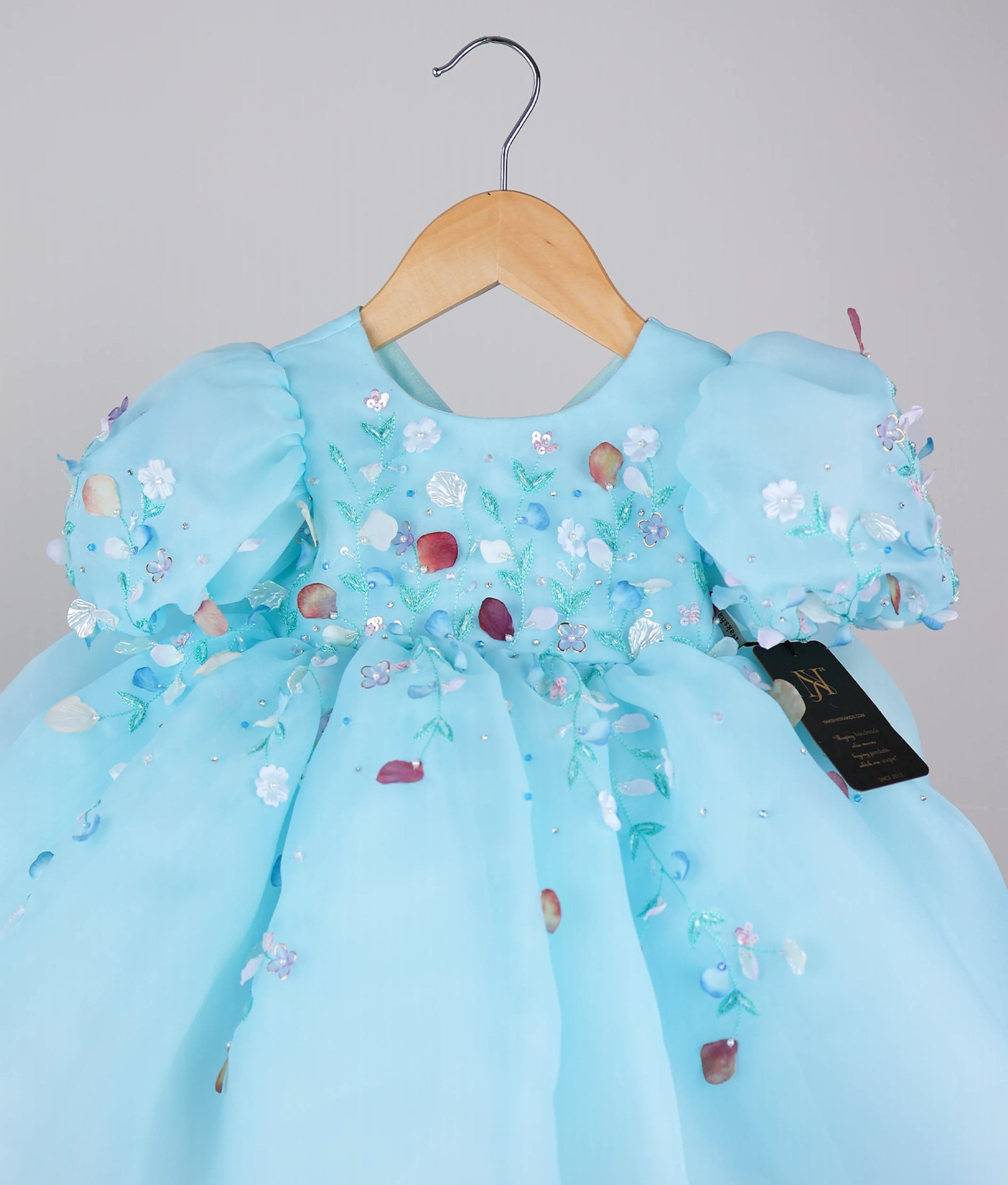 Close view of an blue frock with puff sleeves and floral handwork, displayed on a hanger.