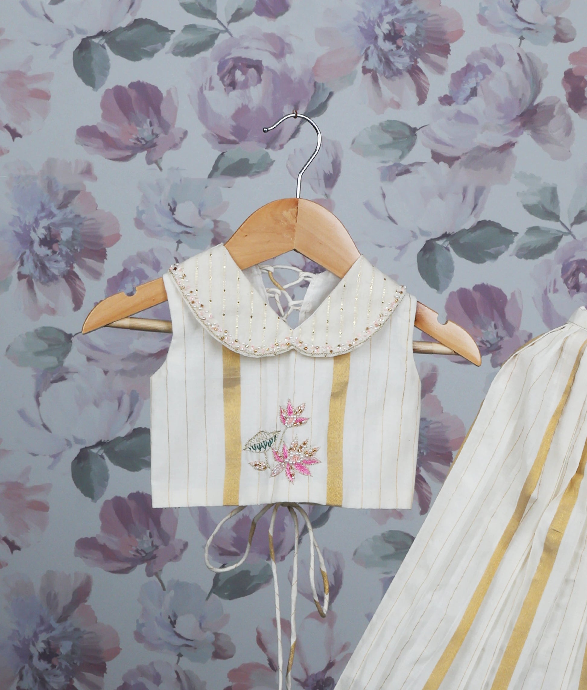 Close view of off-white and gold pleated skirt set with a handworked top on a hanger.