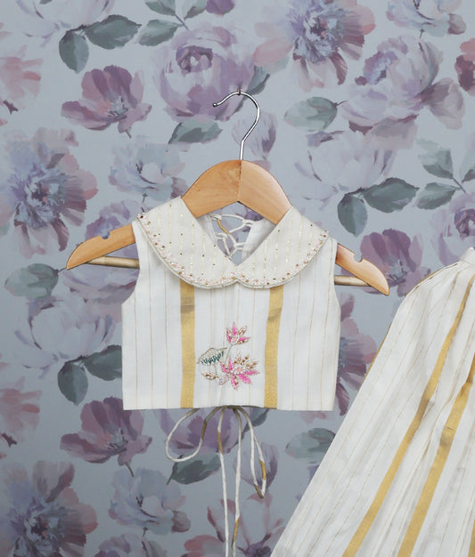 Close view of off-white and gold pleated skirt set with a handworked top on a hanger.