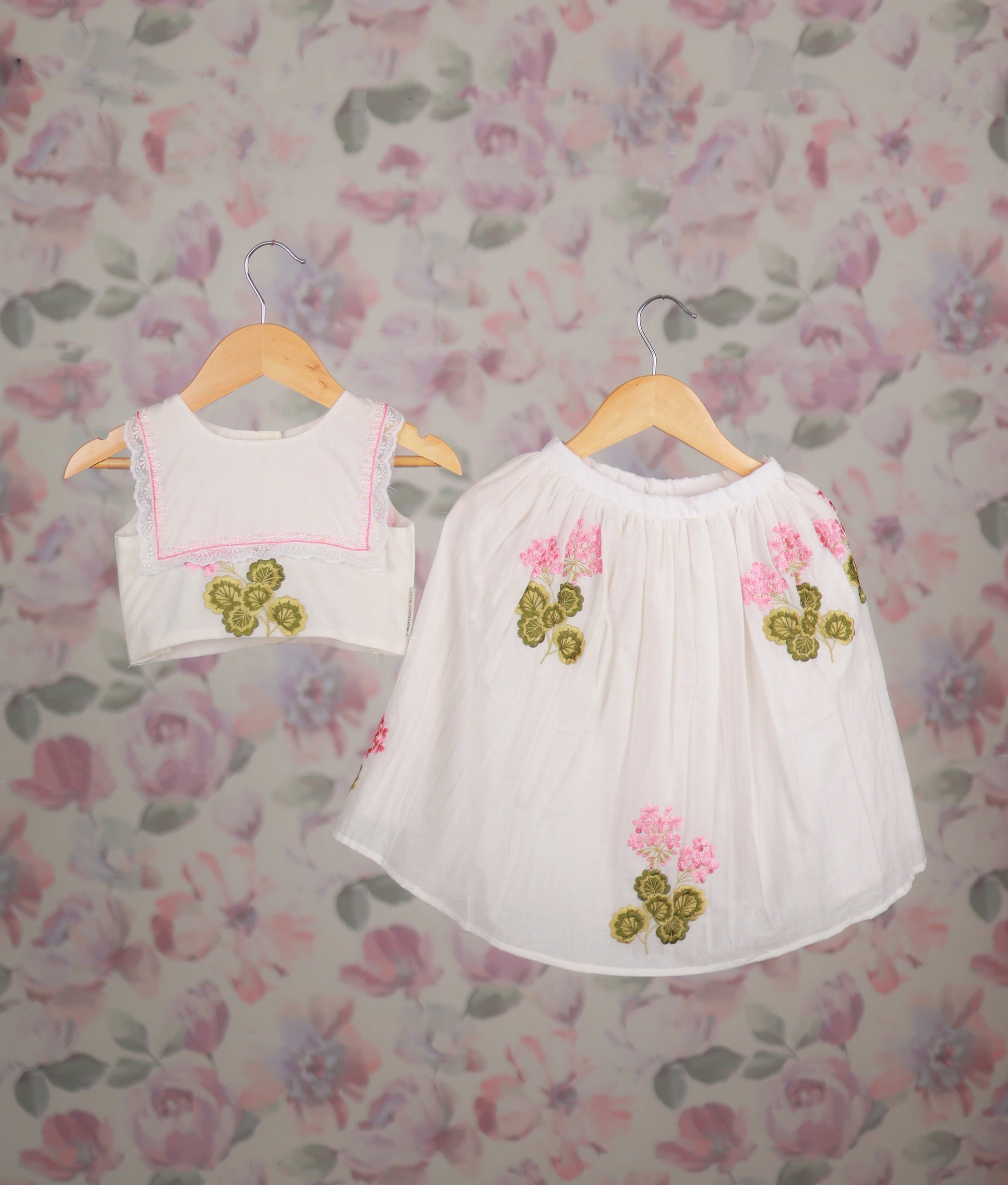 White top and skirt set with pink floral embroidery and lace trim on wooden hangers.