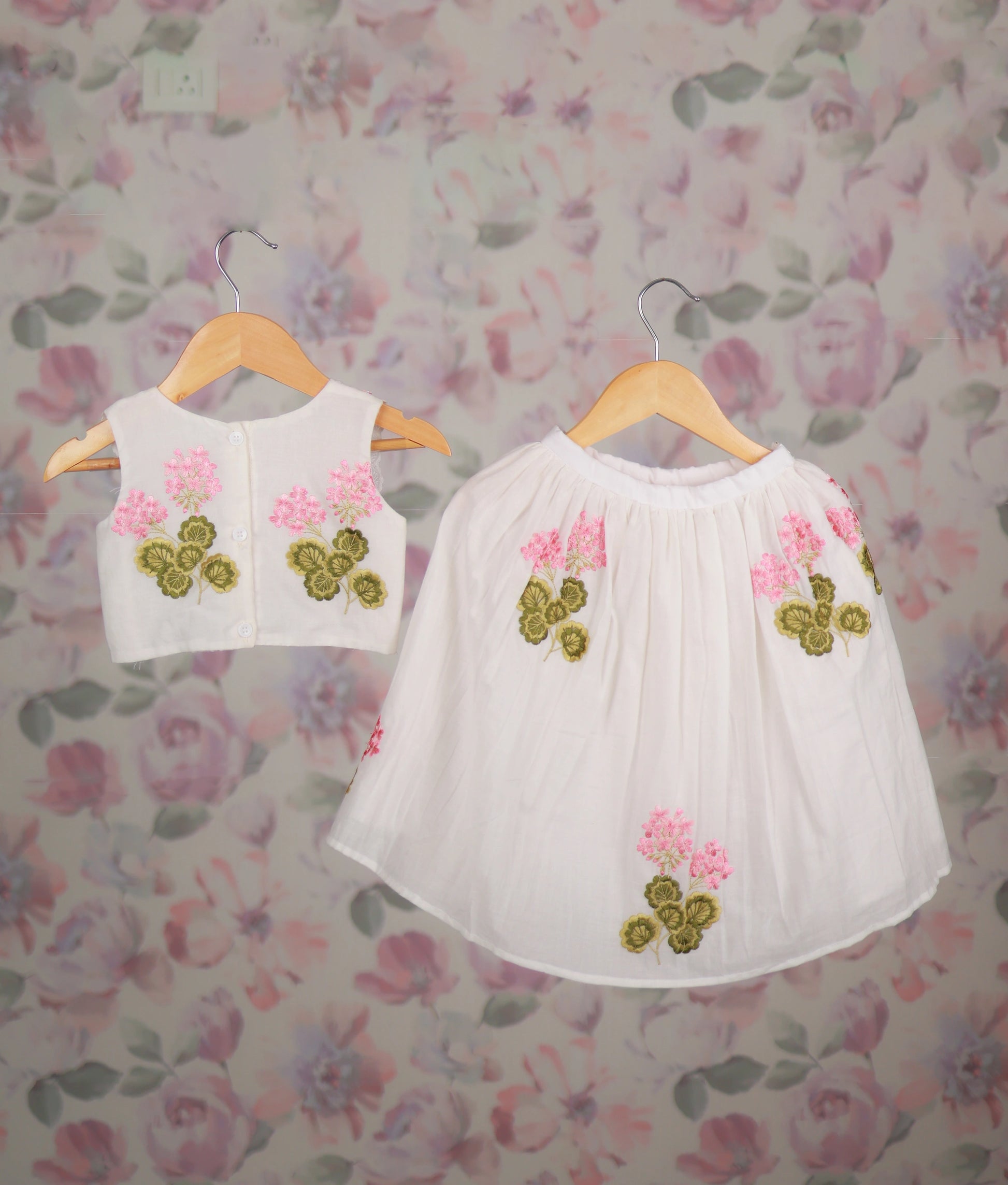 Back view of a white top and skirt set with pink floral embroidery and lace trim on wooden hangers.