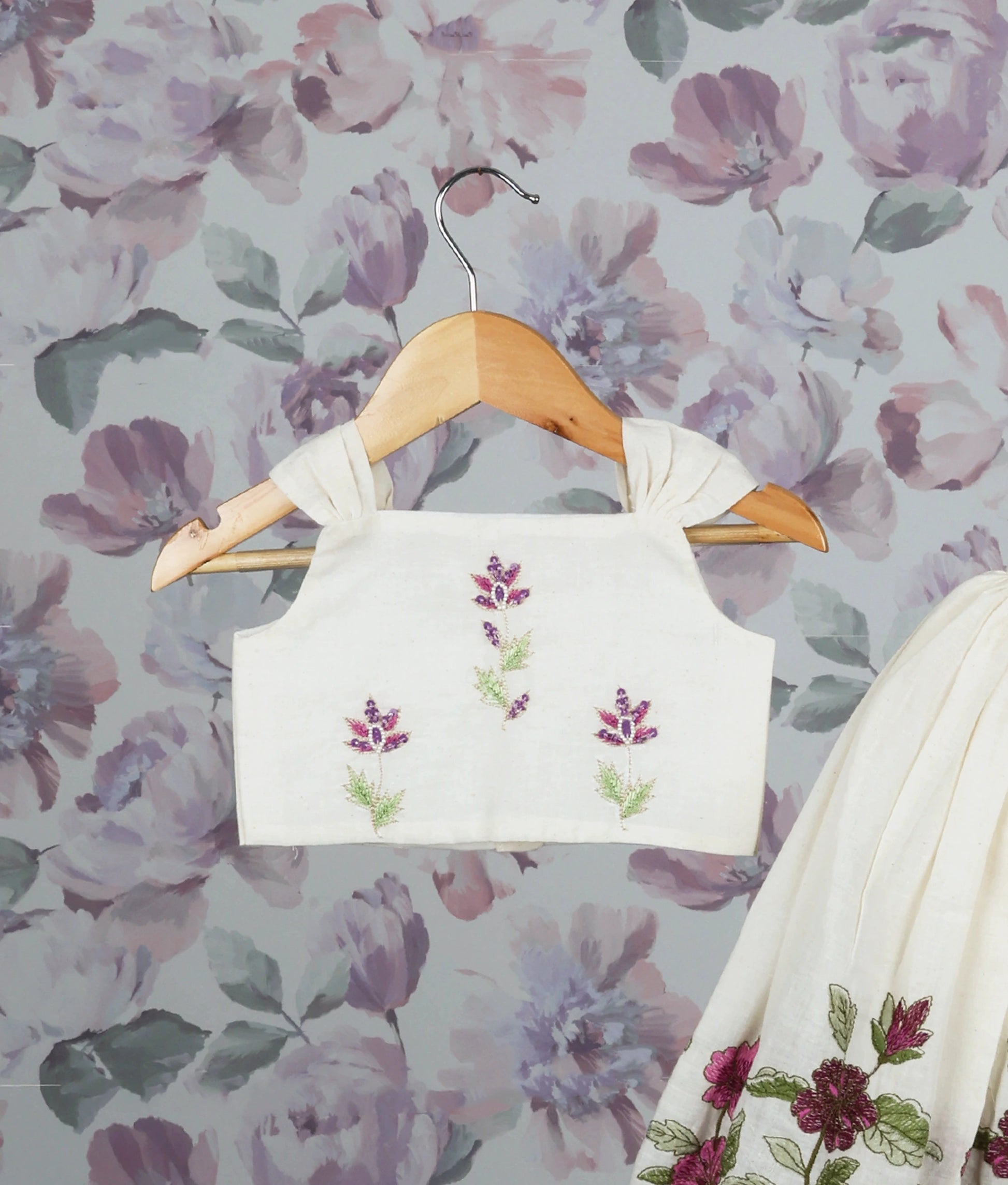 Close view of an floral handworked top and skirt set on hangers against a floral wallpaper background.