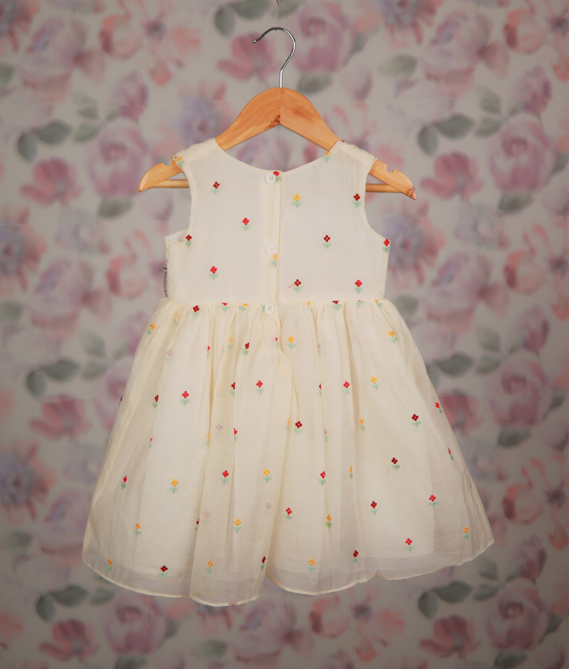 Back view of an off-white frock with colorful floral embroidery against a floral background.