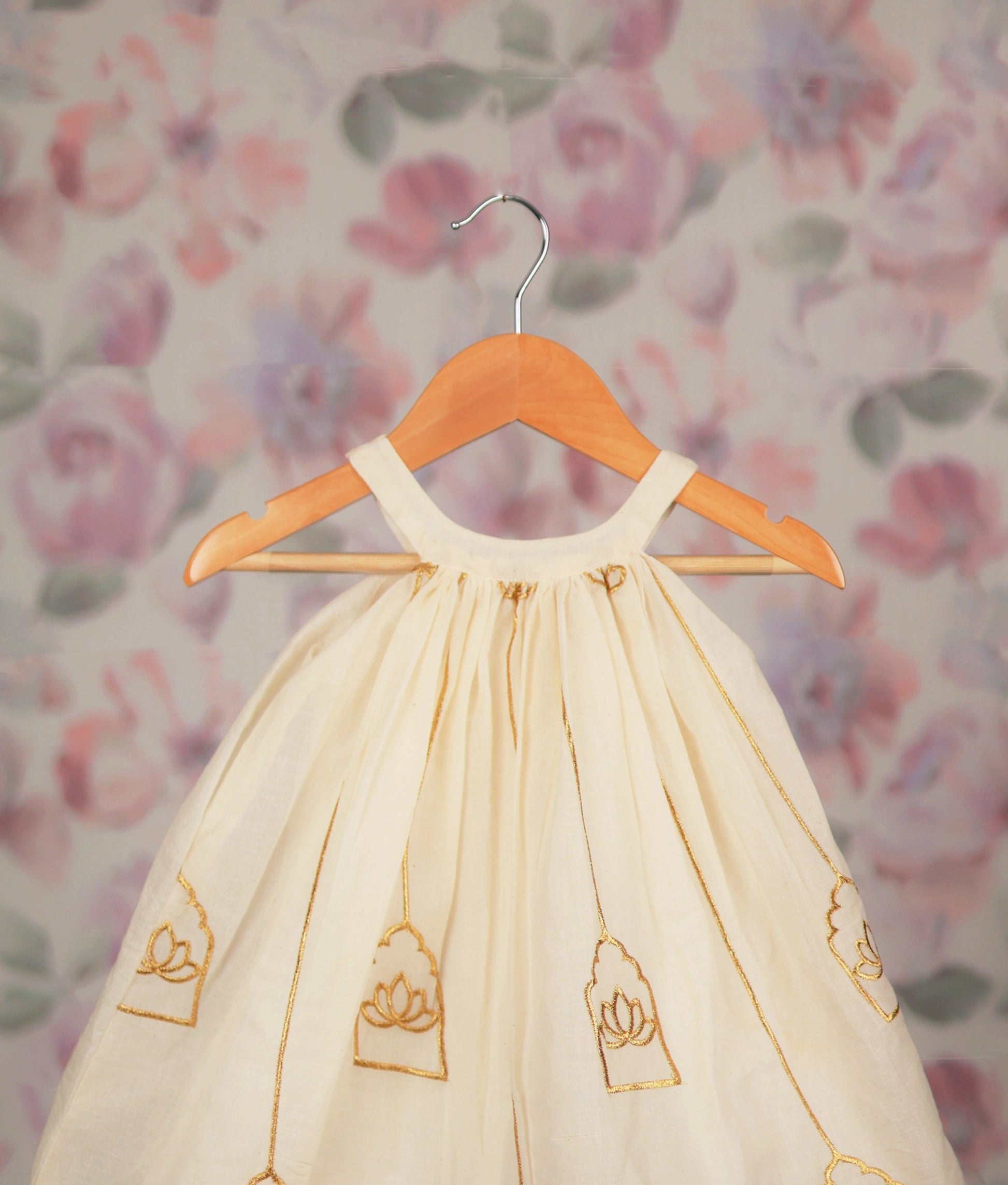 Close view of an off-white gold lotus embroidery frock on a wooden hanger against a floral background.