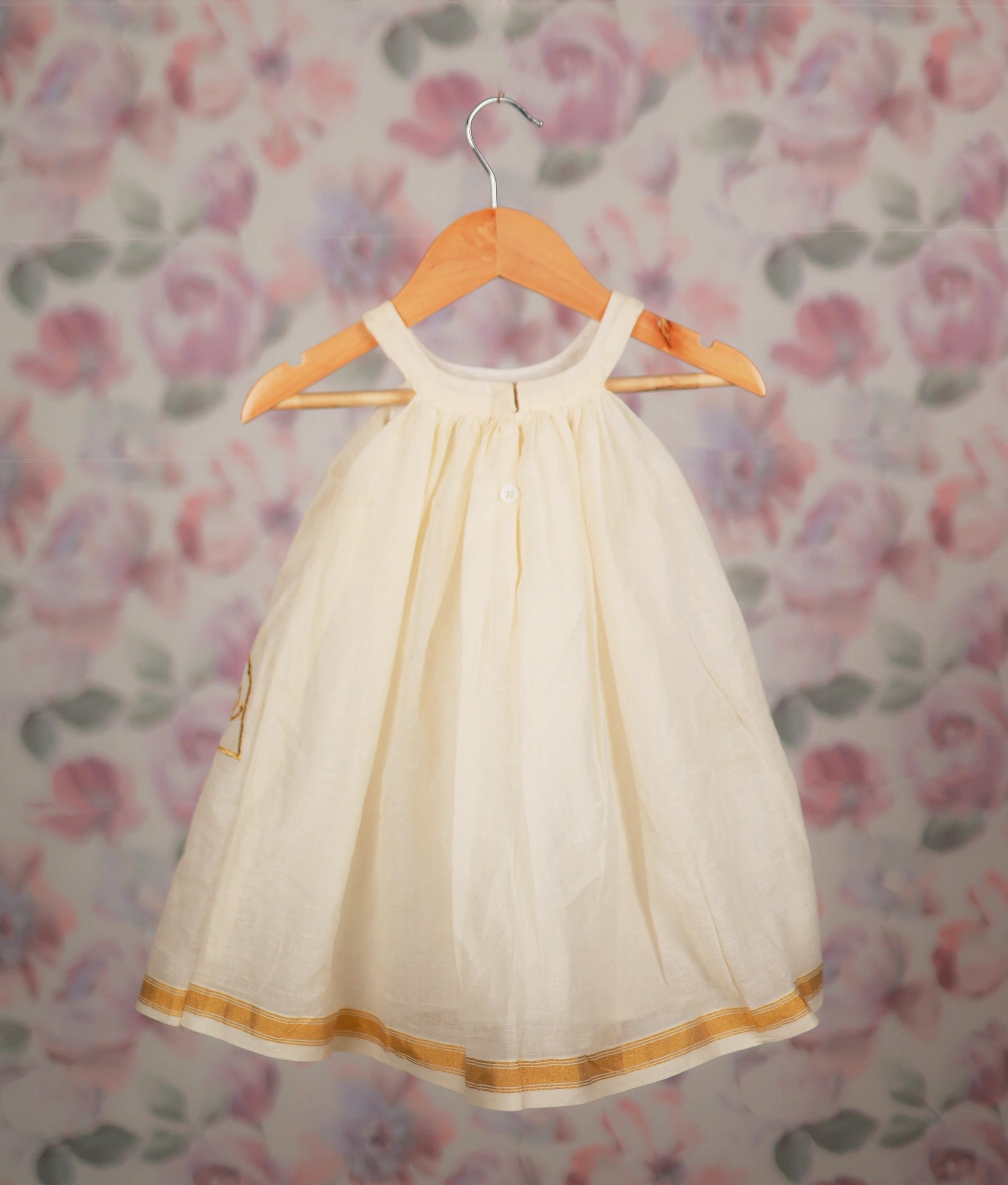 Back view of an off-white gold lotus embroidery frock on a wooden hanger against a floral background.