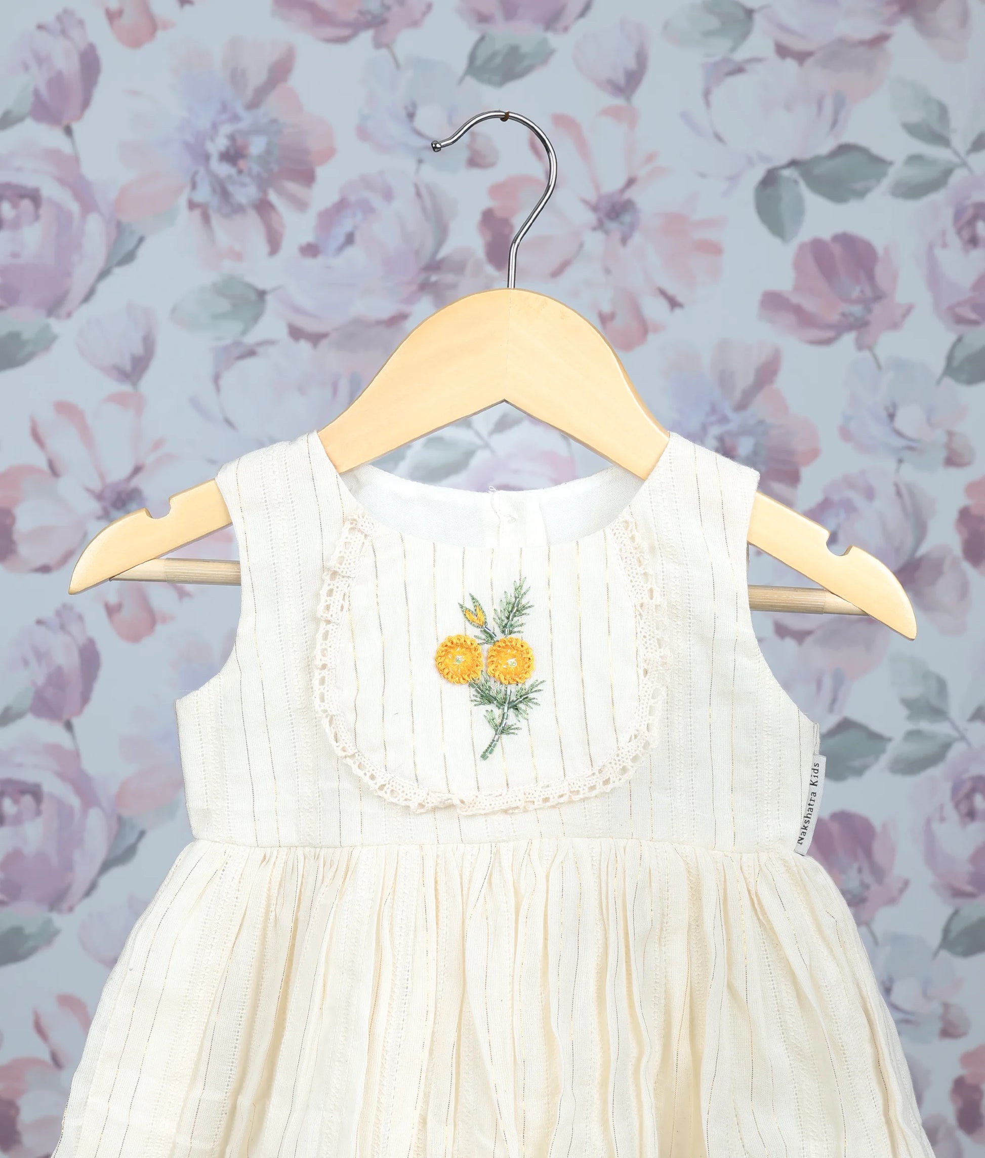 Close view of an off-white sleeveless baby dress with yellow floral embroidery and pleated frock on hangers.