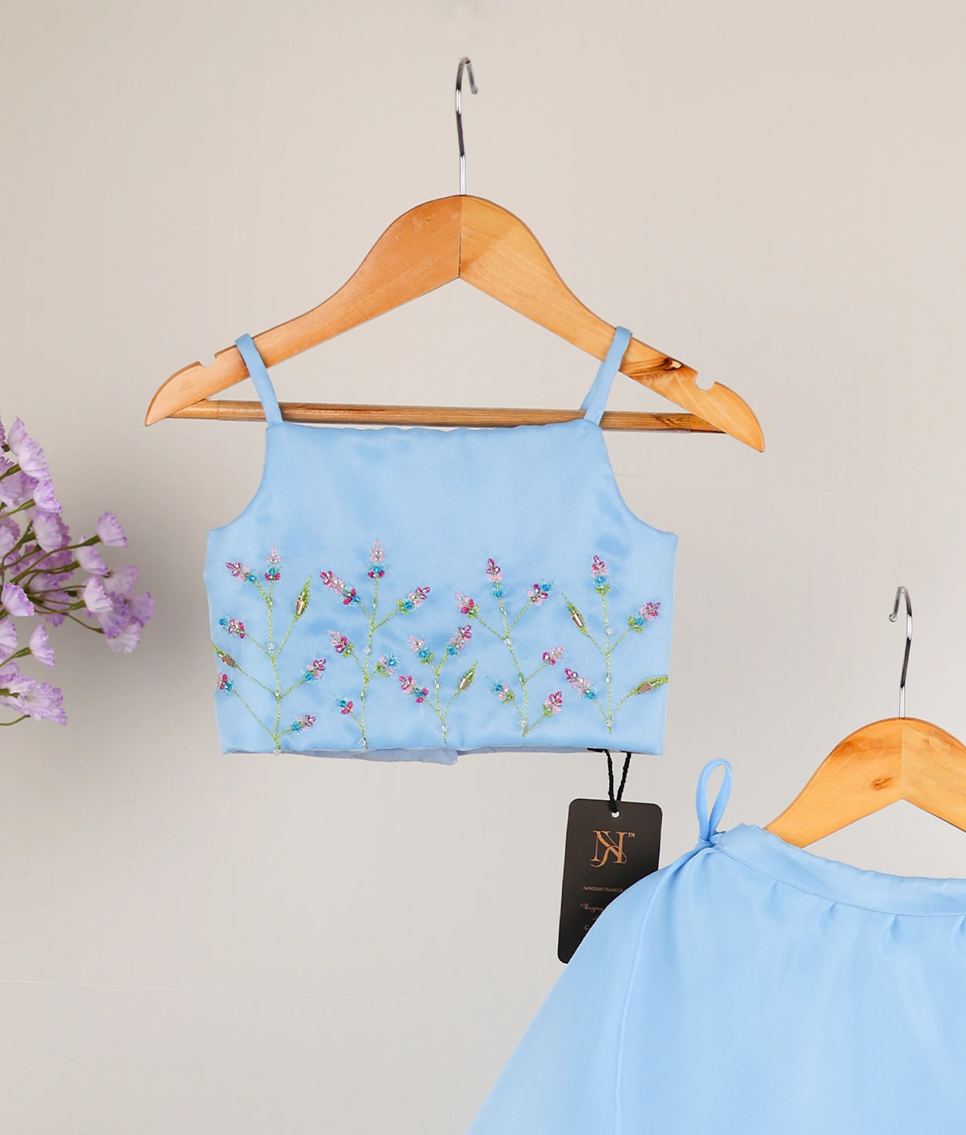 Close view of a sky blue girl's outfit with a handworked floral short top and skirt on wooden hangers.