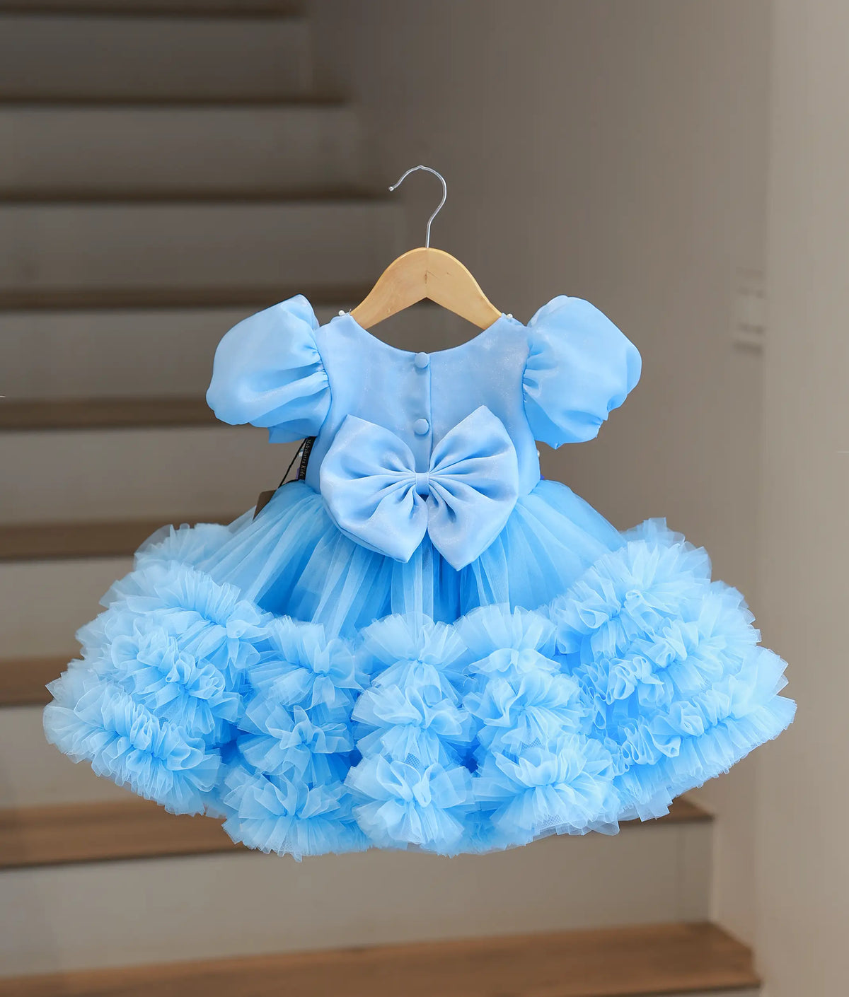 A light blue toddler girl's party dress with puff sleeves, a large bow at the back, and a ruffled tulle skirt, hanging on a wooden hanger against a blurred indoor staircase background.