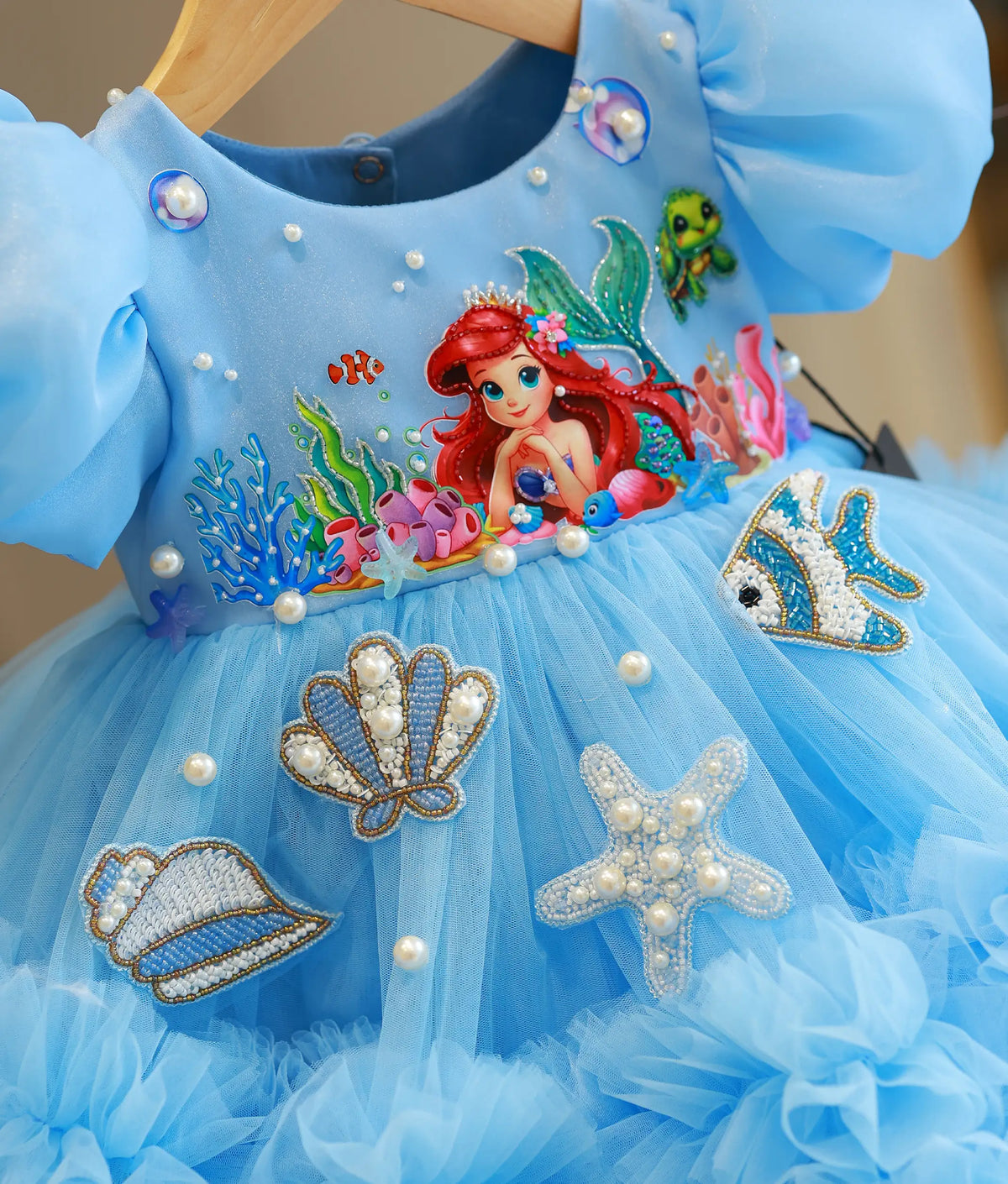 A light blue children's party dress with a tulle skirt, featuring an Ariel-themed underarm patch and appliqués of seashells, a starfish, a fish, and pearl embellishments.