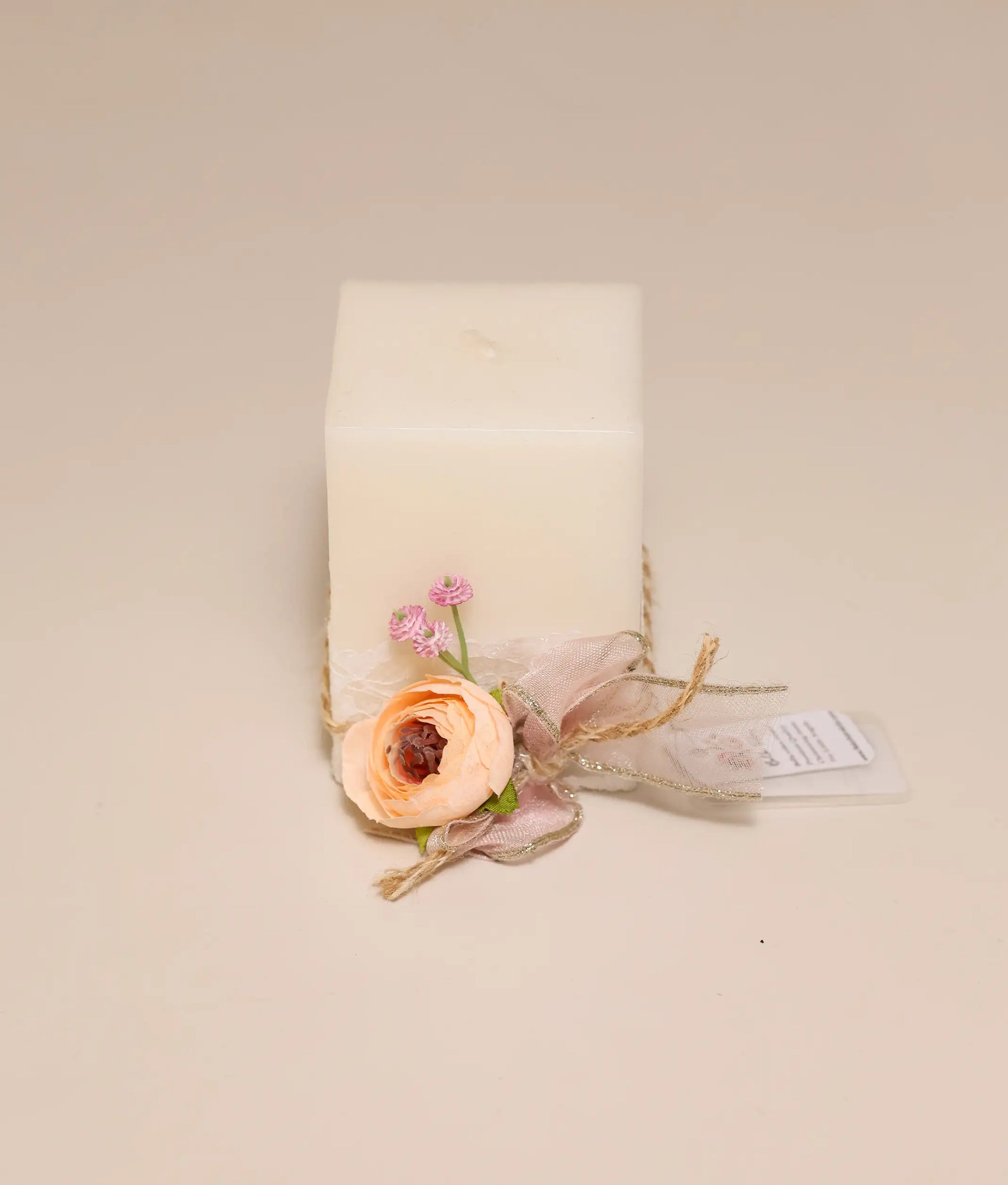Close view of an off-white square candle with a decorative peach rose, pink sprigs, and a dusty rose ribbon. Perfect for your baby's baptism