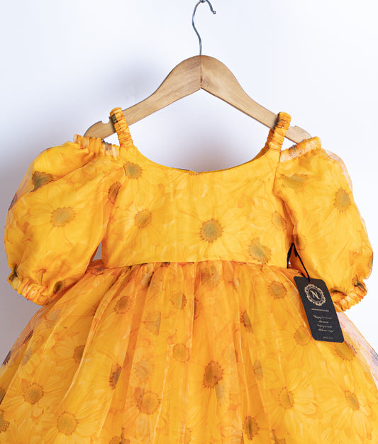 Close view of an yellow printed organza frock with puffed sleeves, displayed on a hanger against a white background.