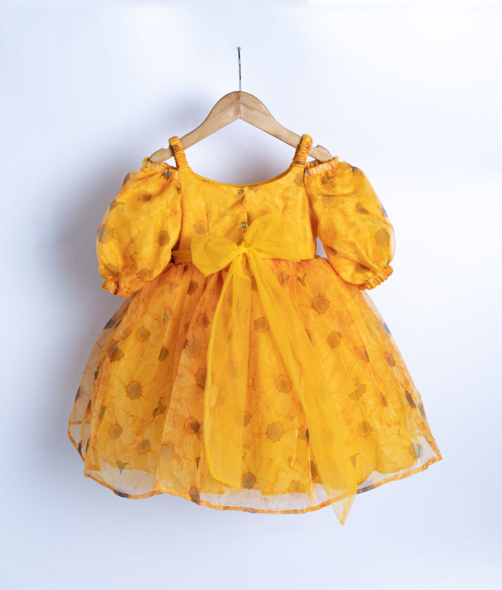 Back view of an yellow printed organza frock with puffed sleeves, displayed on a hanger against a white background.