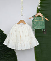Traditional Indian ethnic wear featuring a white embroidered skirt with leaf motifs and a green block-printed blouse with golden detailing, displayed on wooden hangers with a tropical leaf backdrop.