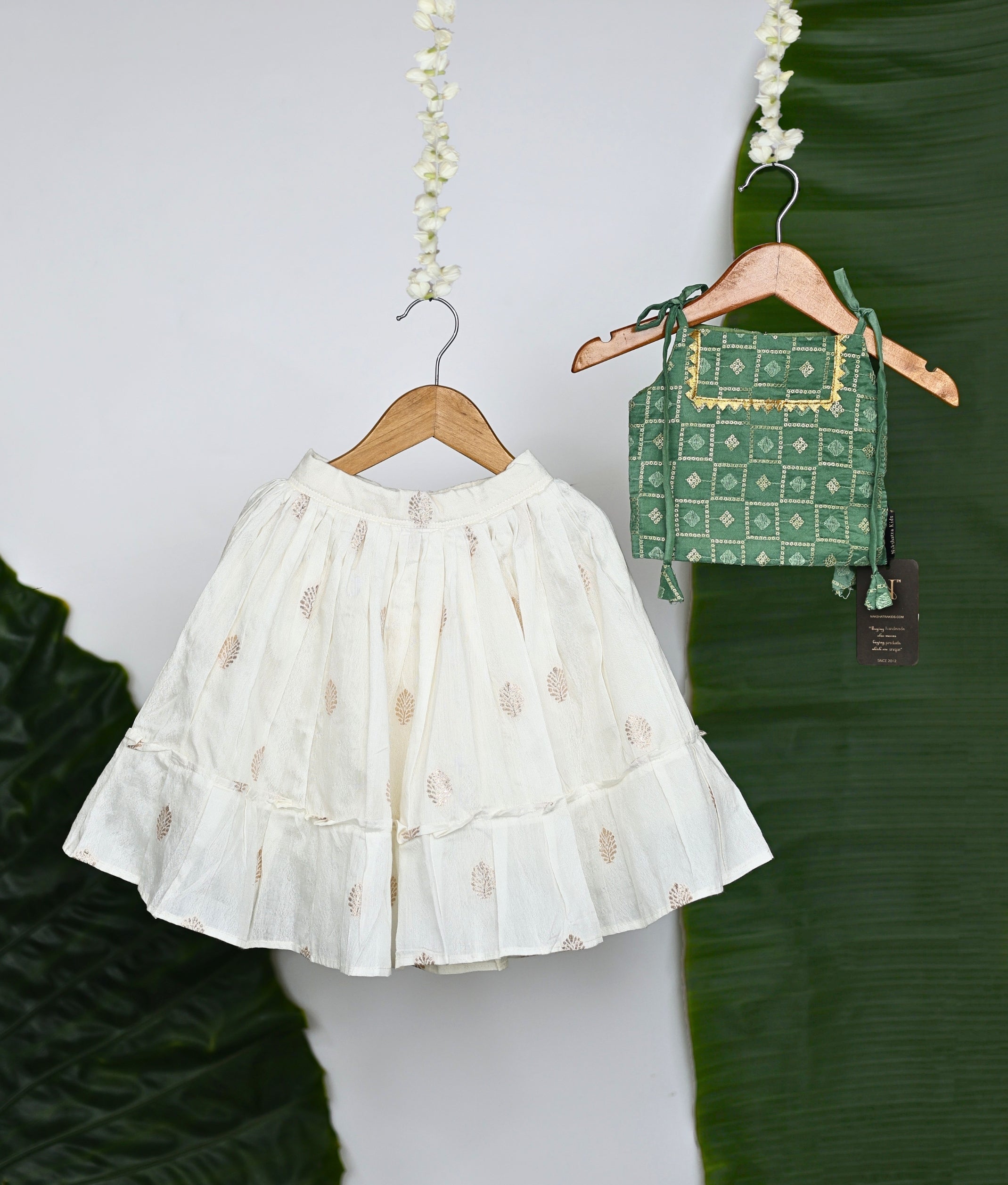 Traditional Indian ethnic wear featuring a white embroidered skirt with leaf motifs and a green block-printed blouse with golden detailing, displayed on wooden hangers with a tropical leaf backdrop.