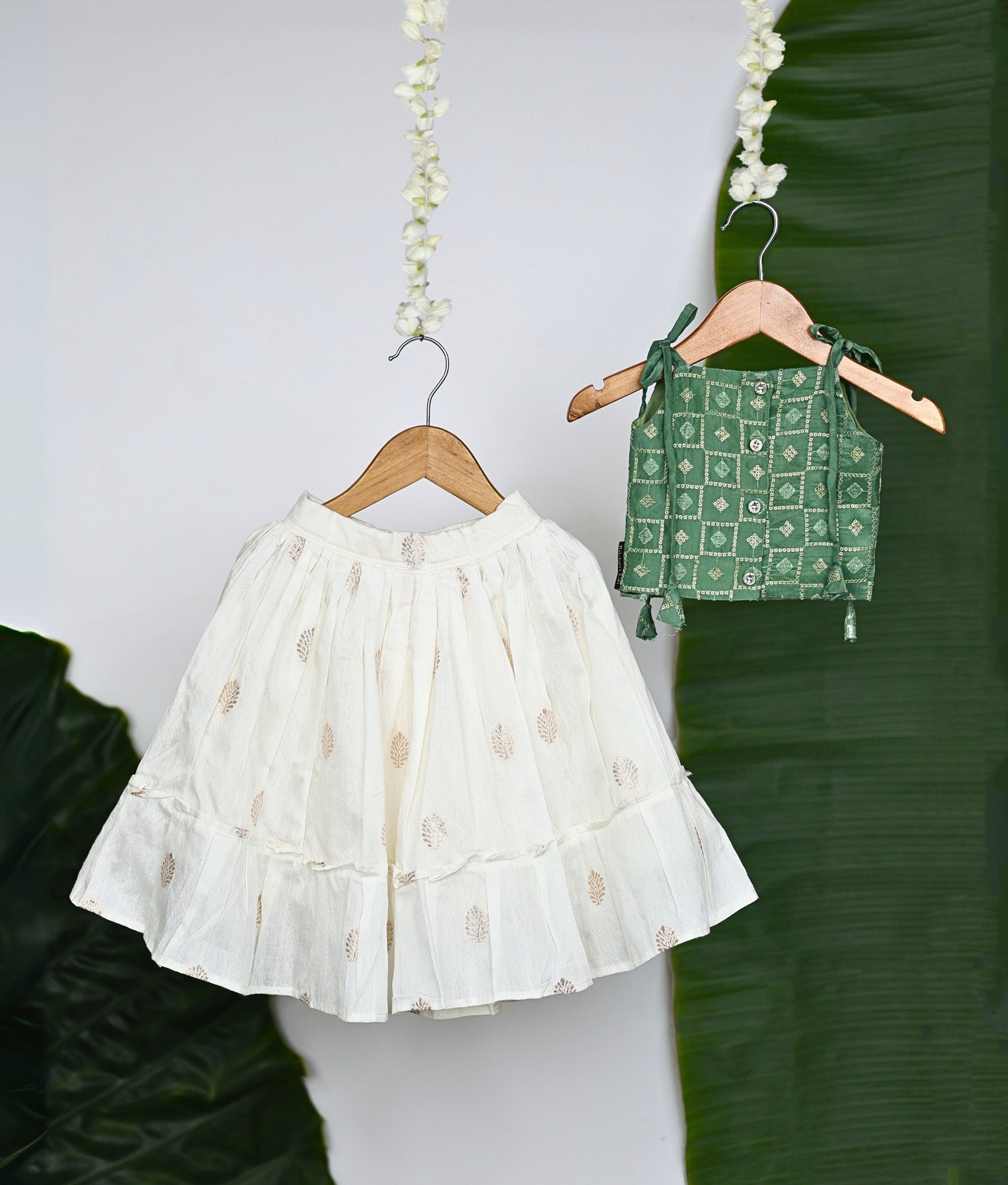 Back view of traditional Indian ethnic wear for girls featuring a white embroidered skirt with leaf motifs and a green block-printed blouse with golden detailing, displayed on wooden hangers with a tropical leaf backdrop.