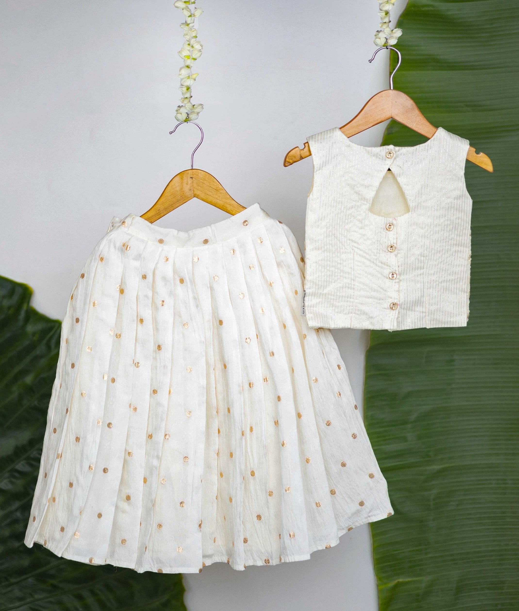 Back view of a traditional white and gold sleeveless blouse with embroidered detailing paired with a white pleated skirt featuring gold polka dots—a festive ethnic outfit for girls displayed on hangers against a leafy background.
