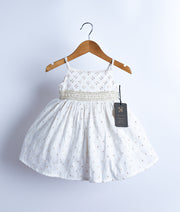 An off-white, sleeveless, flared frock with a band at the waist, displayed on a wooden hanger against a white background.