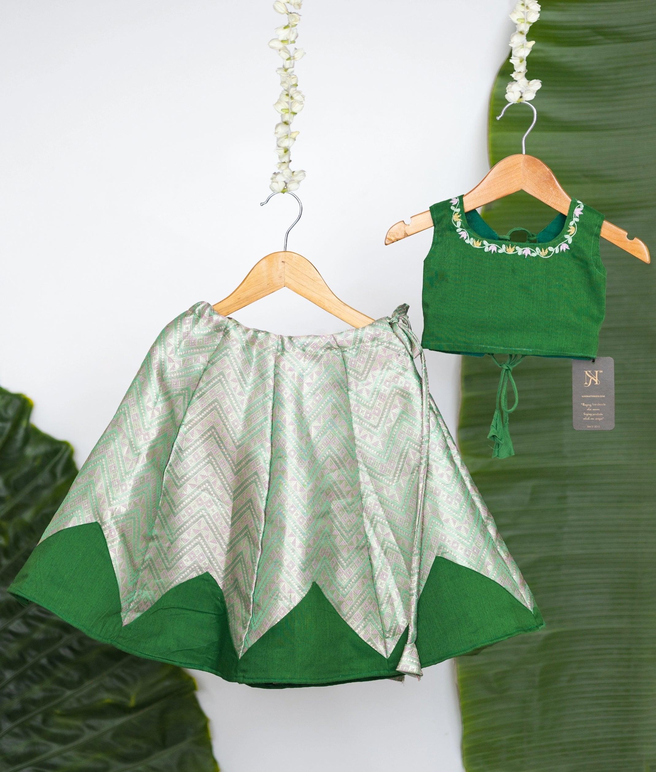 Stylish green kids set with embroidered sleeveless blouse and chevron-patterned skirt, displayed on wooden hangers against a leaf backdrop.