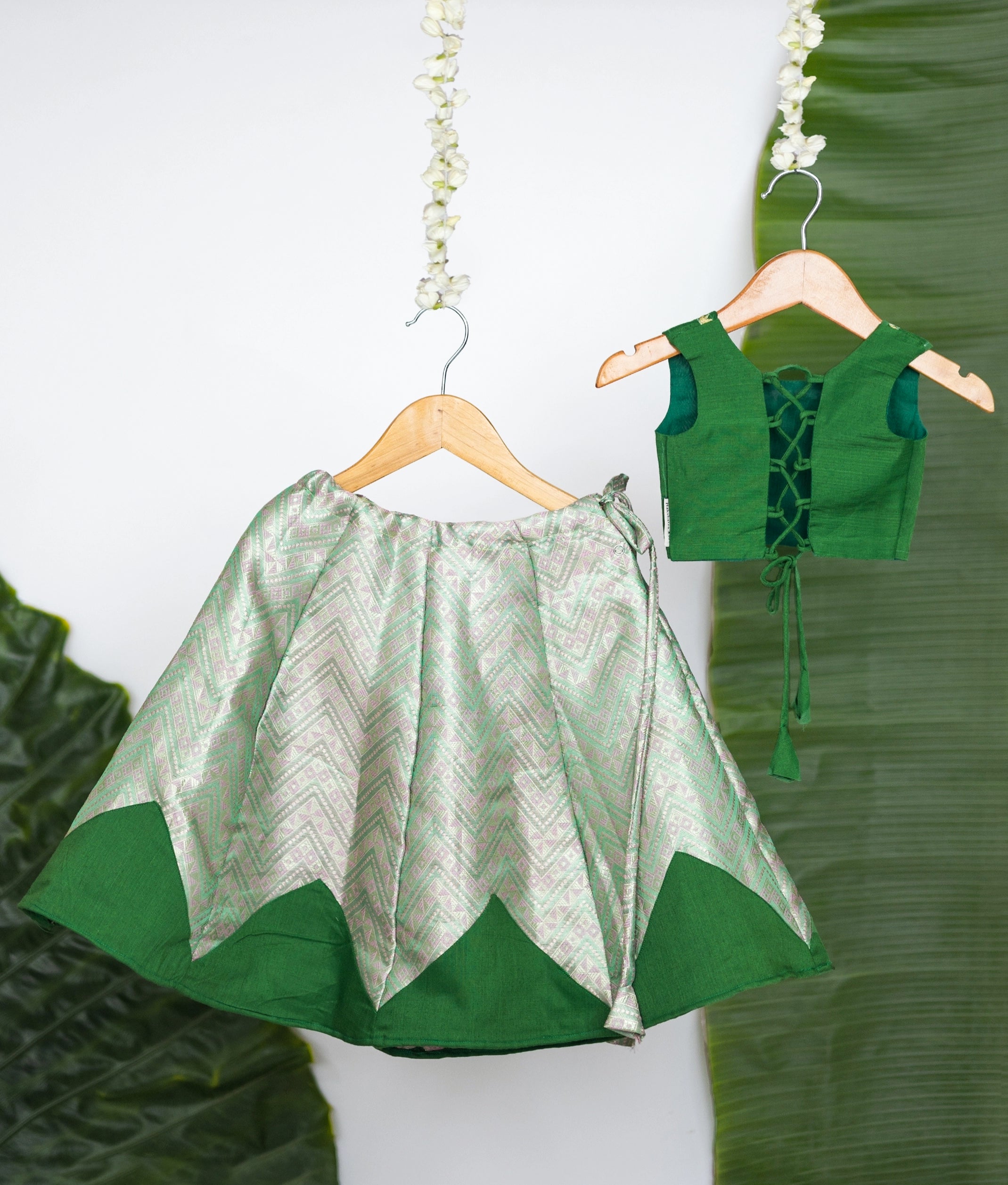 Back view of a stylish green kids set with an embroidered sleeveless blouse and chevron-patterned skirt, displayed on wooden hangers against a leaf backdrop.