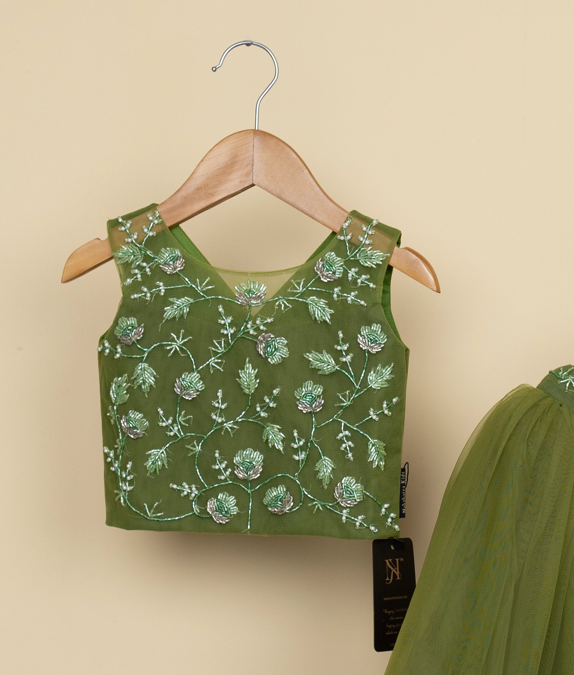 Close view of a olive green floral tulle net top and skirt on hangers against a beige wall background.
