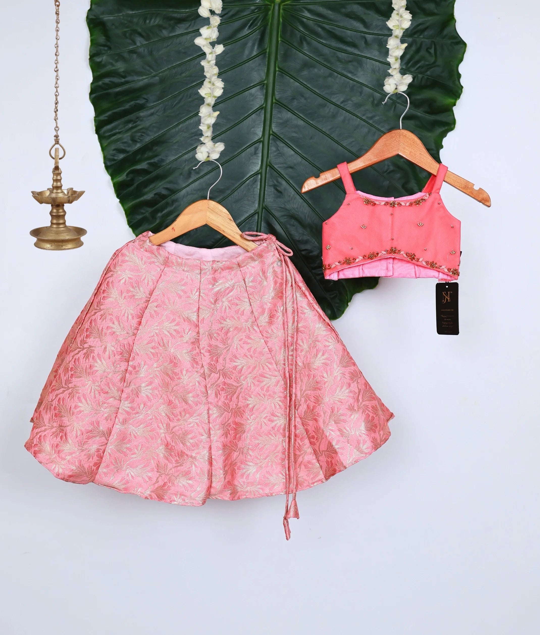 Stylish pink kids lehenga set with floral brocade skirt and embellished sleeveless top, displayed on hangers against a large green leaf backdrop with decorative brass lamp.
