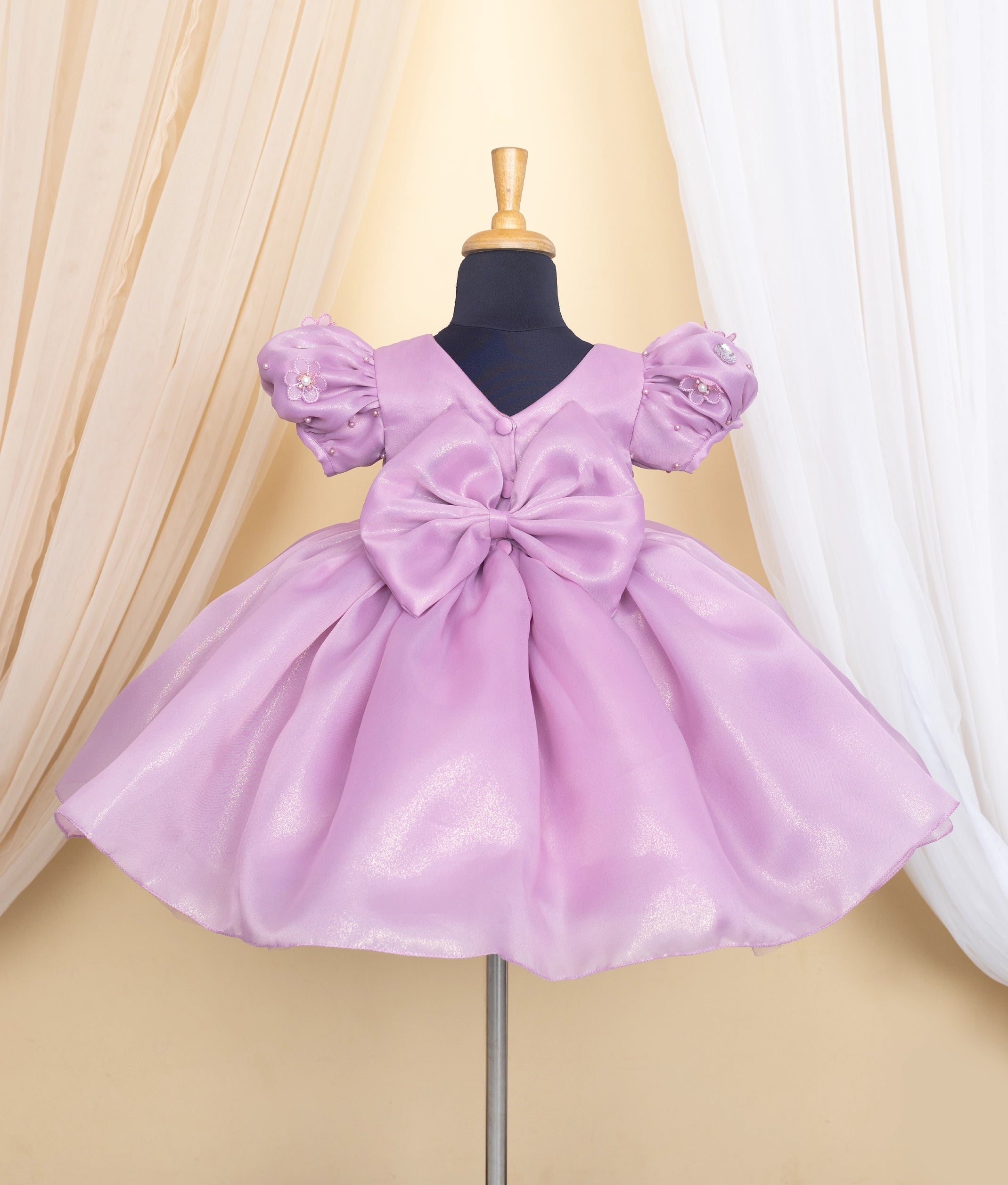 Back view of an lavender frock on a mannequin against a beige curtain background.
