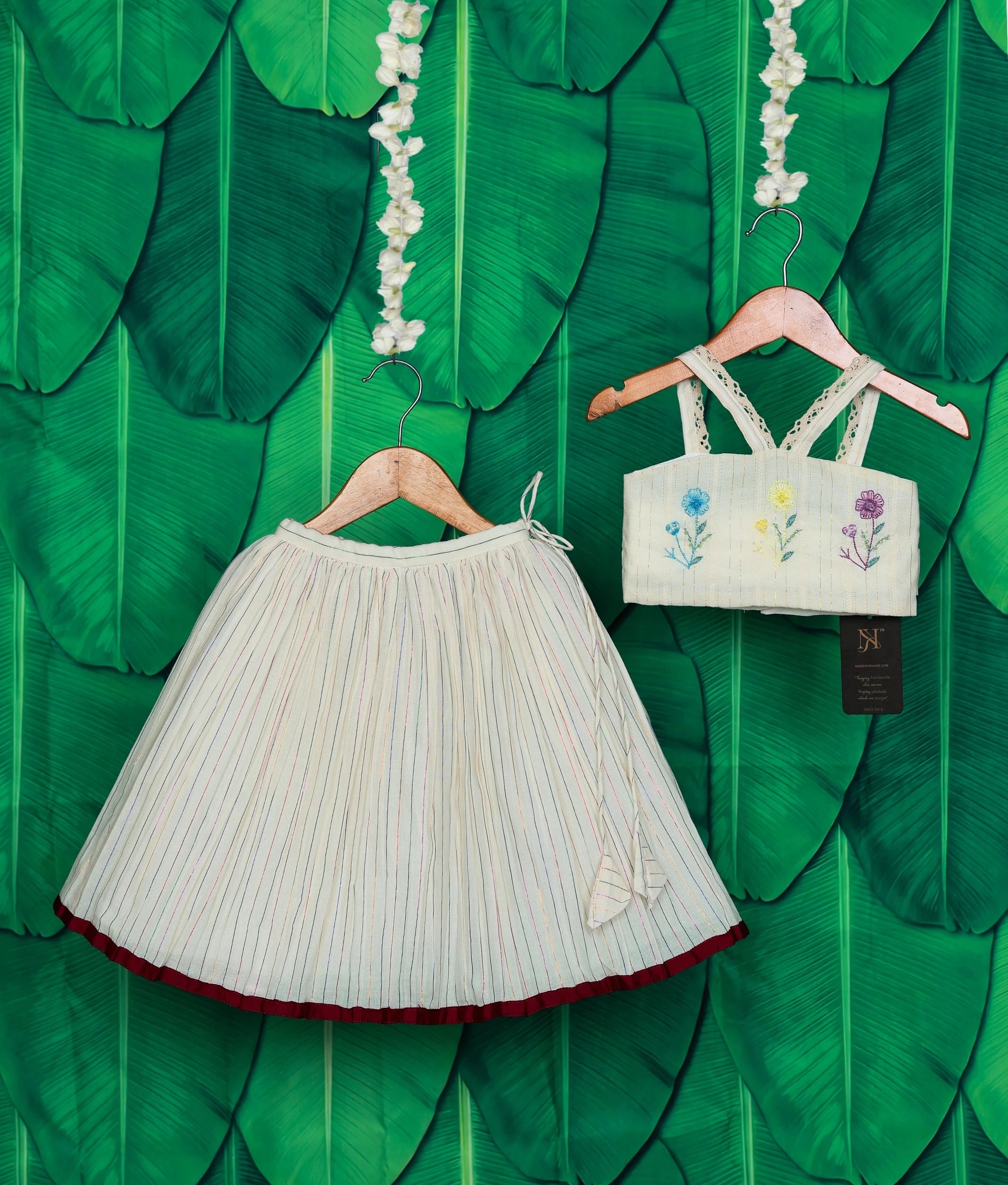 An off-white skirt with a maroon hem and a floral embroidery top on a green leaf background.
