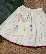 Off-white skirt with a maroon hem and a floral embroidery top on a green leaf and a woven mat background.