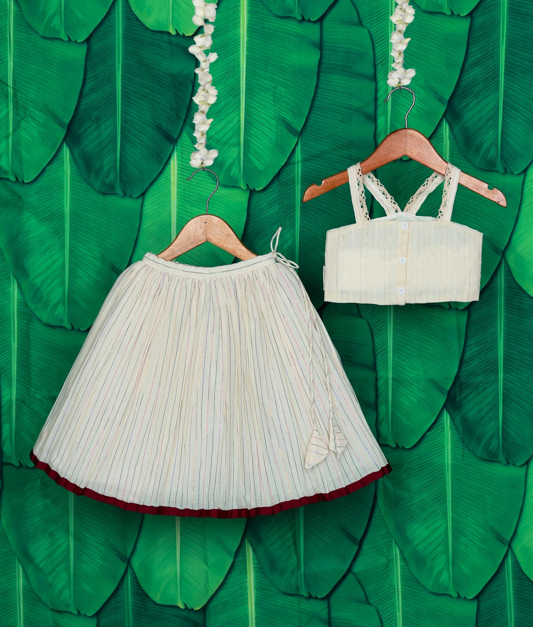 Back view of an off-white skirt with a maroon hem and sleeveless top set with lace straps, displayed on a green leaf backdrop.