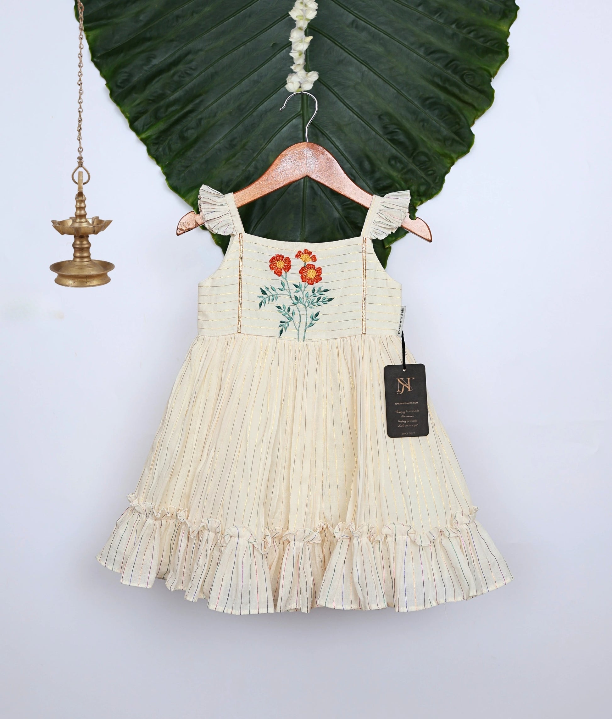 An off-white pleated frock with floral embroidery and ruffled sleeves and hem, displayed on a hanger with decor.