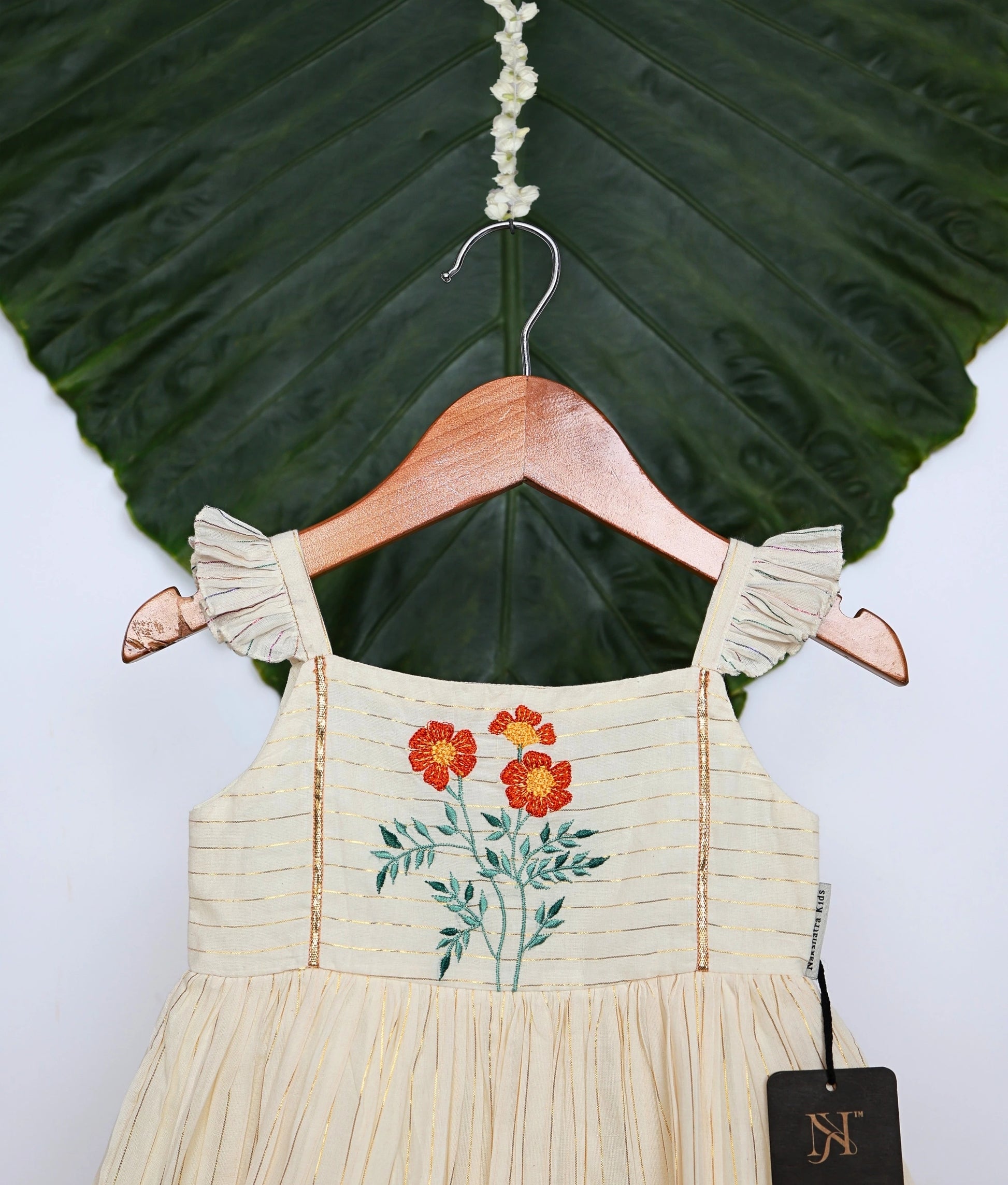 Close view of an off-white pleated frock with floral embroidery and ruffled sleeves and hem, displayed on a hanger.