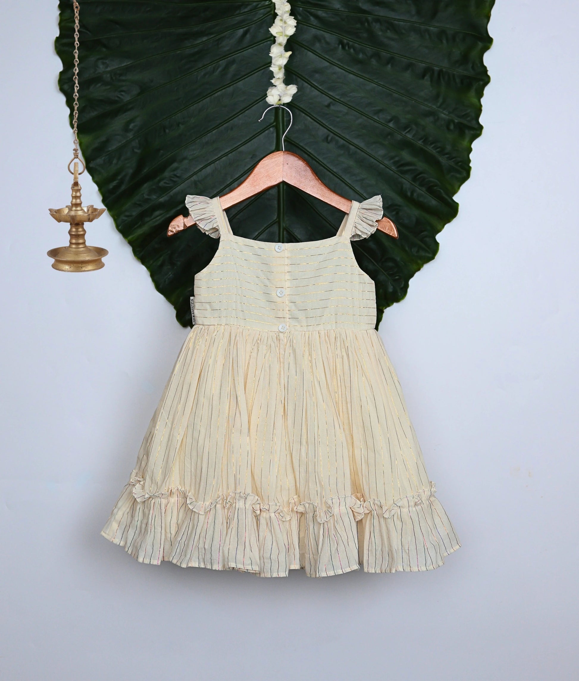 Back view of a pleated frock with floral embroidery and ruffled sleeves and hem, displayed on a hanger with decor.