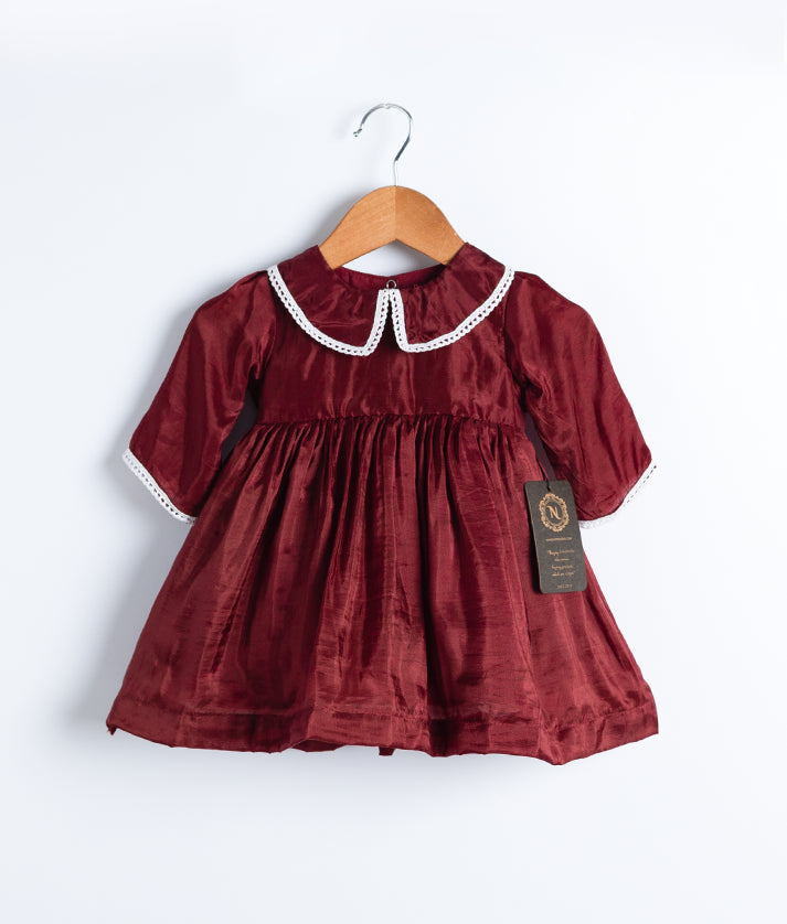 A maroon christmas frock with a yoke and peter pan collar, pleated skirt, and lace detailing, displayed on a hanger.