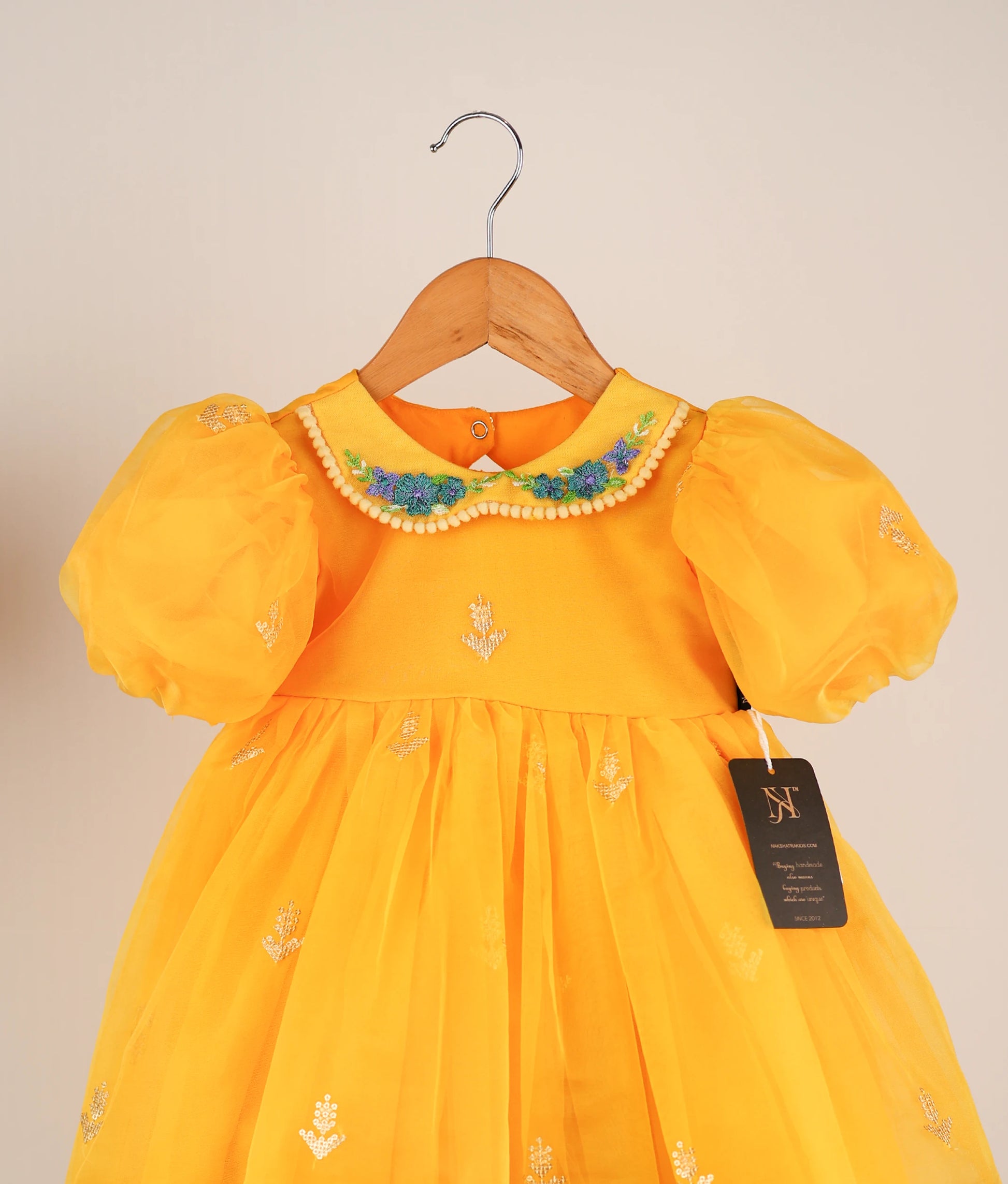 Close view of a yellow organza baby frock with embroidery and a round neckline, displayed on a hanger.