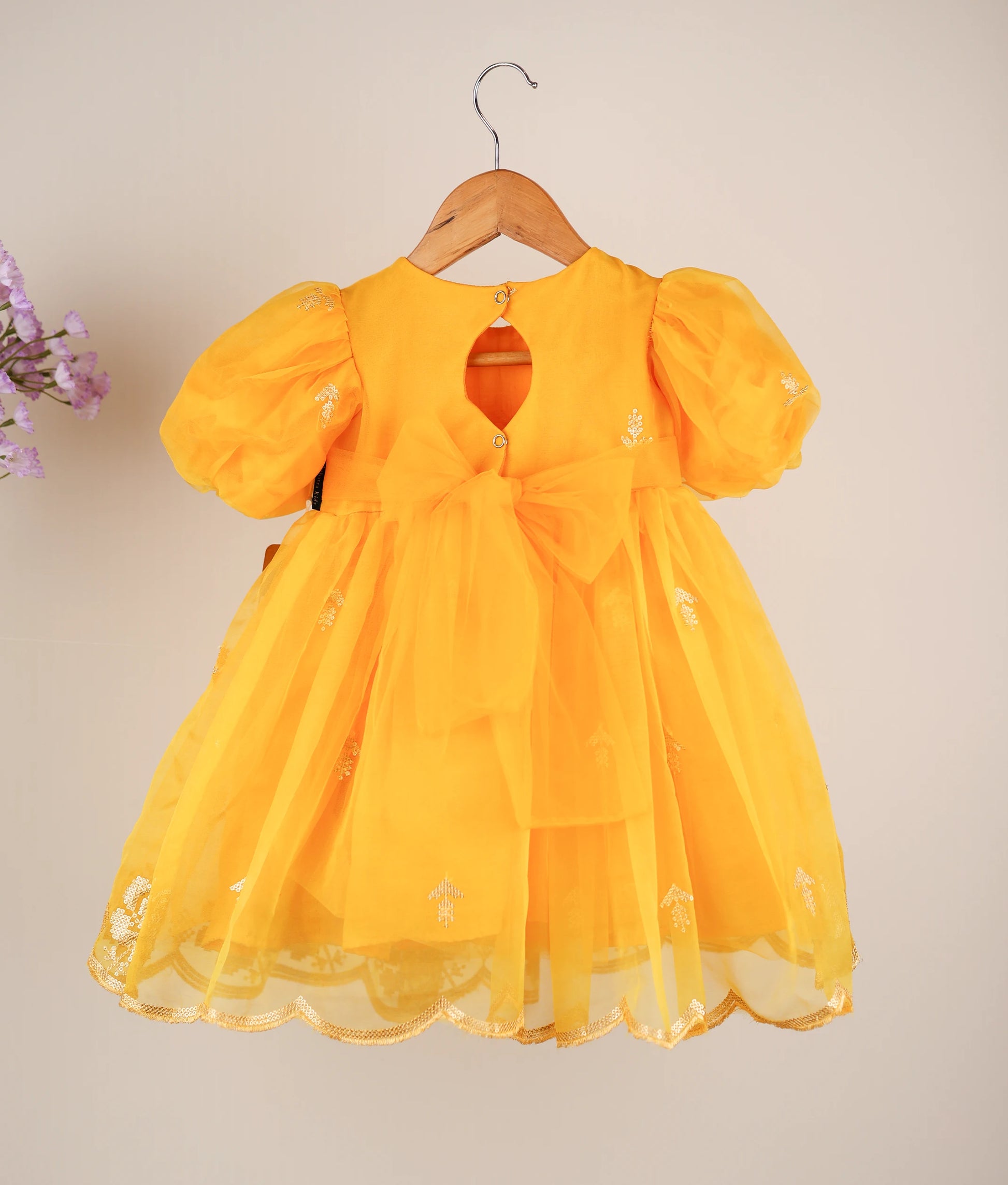 Back view of a yellow organza baby frock with embroidery and a round neckline, displayed on a hanger.