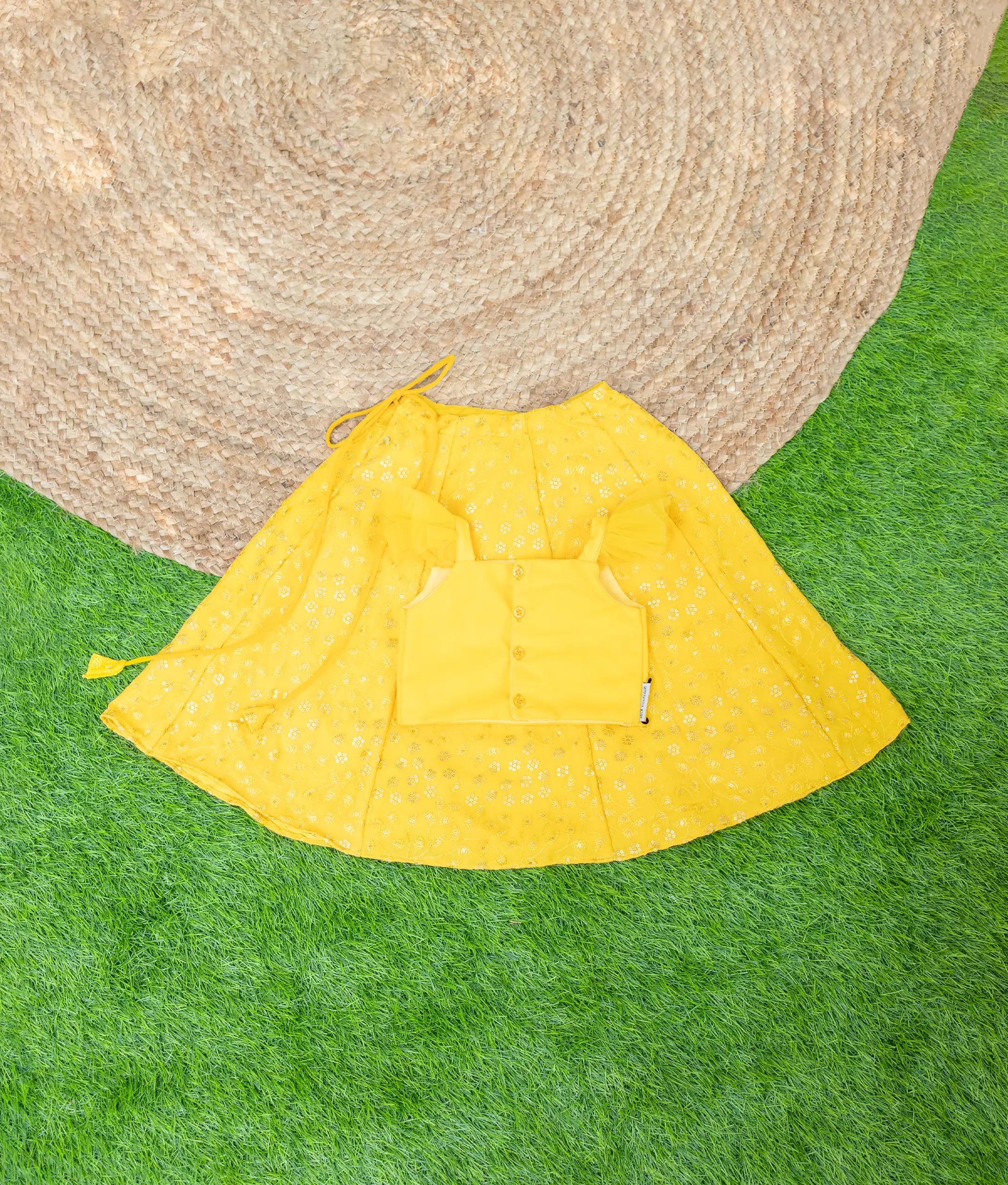 Back view of a girls yellow festive outfit featuring a buttoned crop top with ruffled straps and a matching flared skirt with floral eyelet embroidery, displayed on grass with a jute mat backdrop.