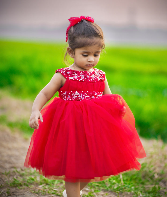 A baby girl wearing a red tulle net handworked birthday frock with embroidery on the neckline and waist, standing against a natural background.