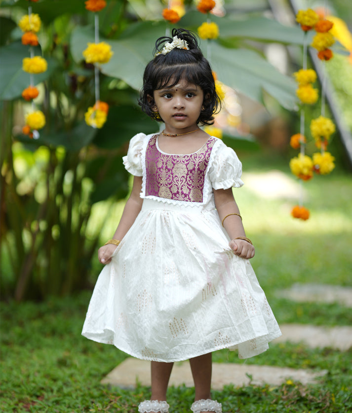 A child wearing an off-white short-sleeved frock with a round neckline, featuring lace details and a flared design, standing in a garden setting.