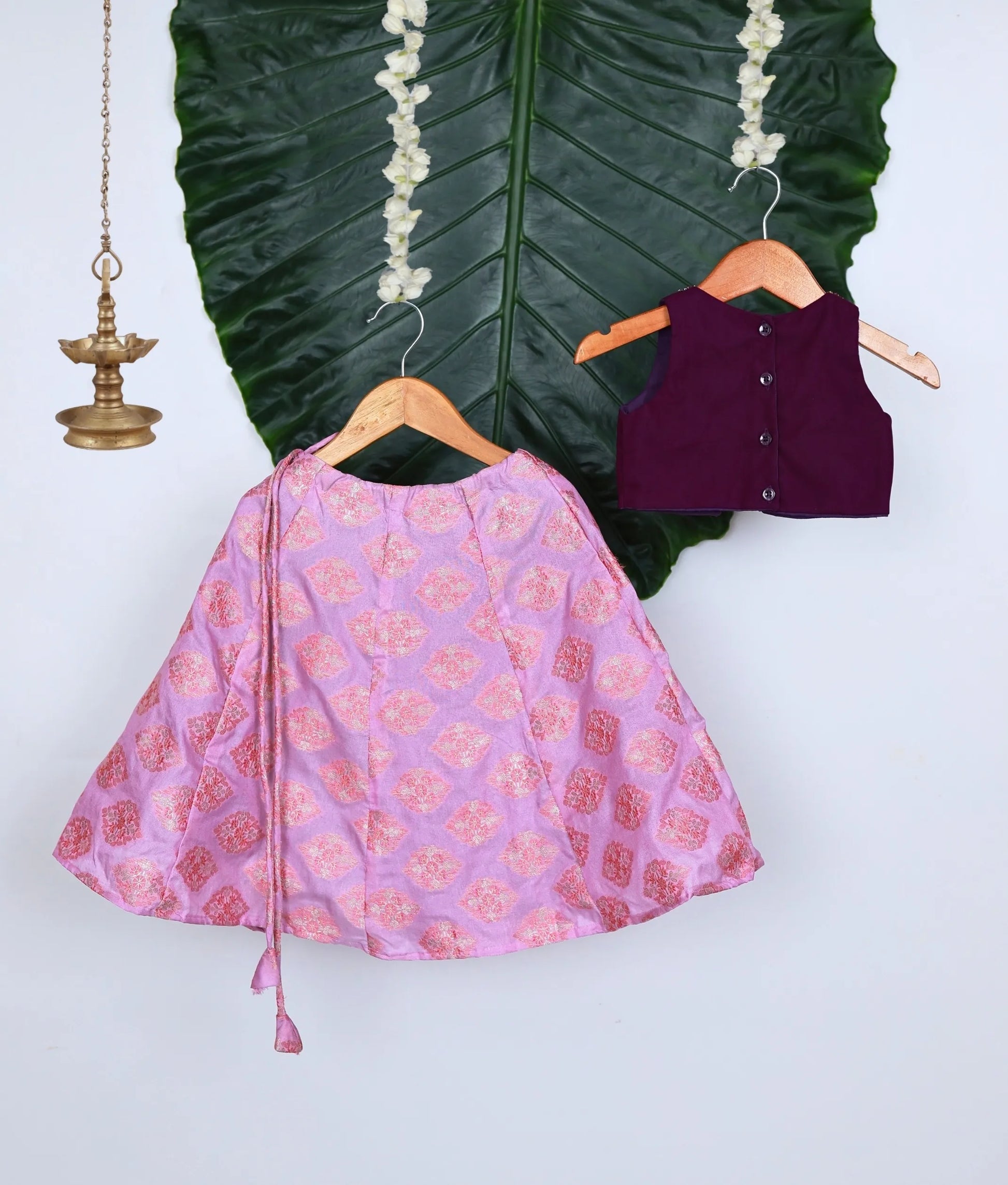 Back view of a traditional kids lehenga set featuring a purple handworked sleeveless blouse and a pink brocade skirt with gold motifs.